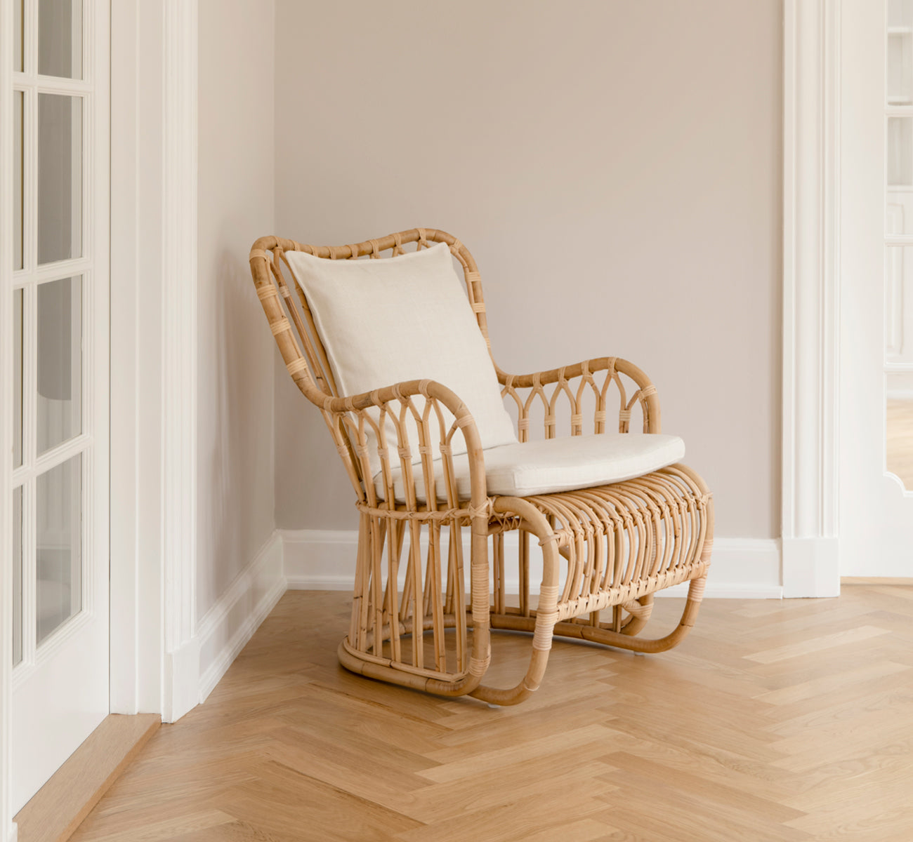 Dette er et livsstilsbillede af Tulip Lænestol i polished natural rattan. Kombineret med en matchende sofa i et lyst panelrum med dør.
