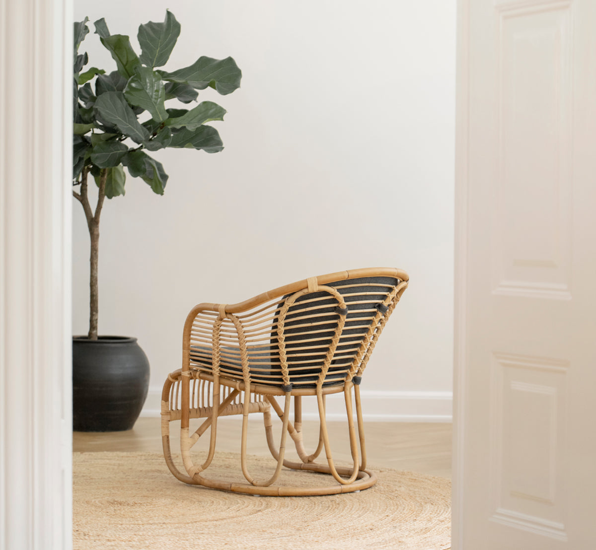 Dette er et livsstilsbillede af Swing Lænestol i polished natural rattan. Stående alene ved siden af en løvrig plante på et sildebensparketgulv.