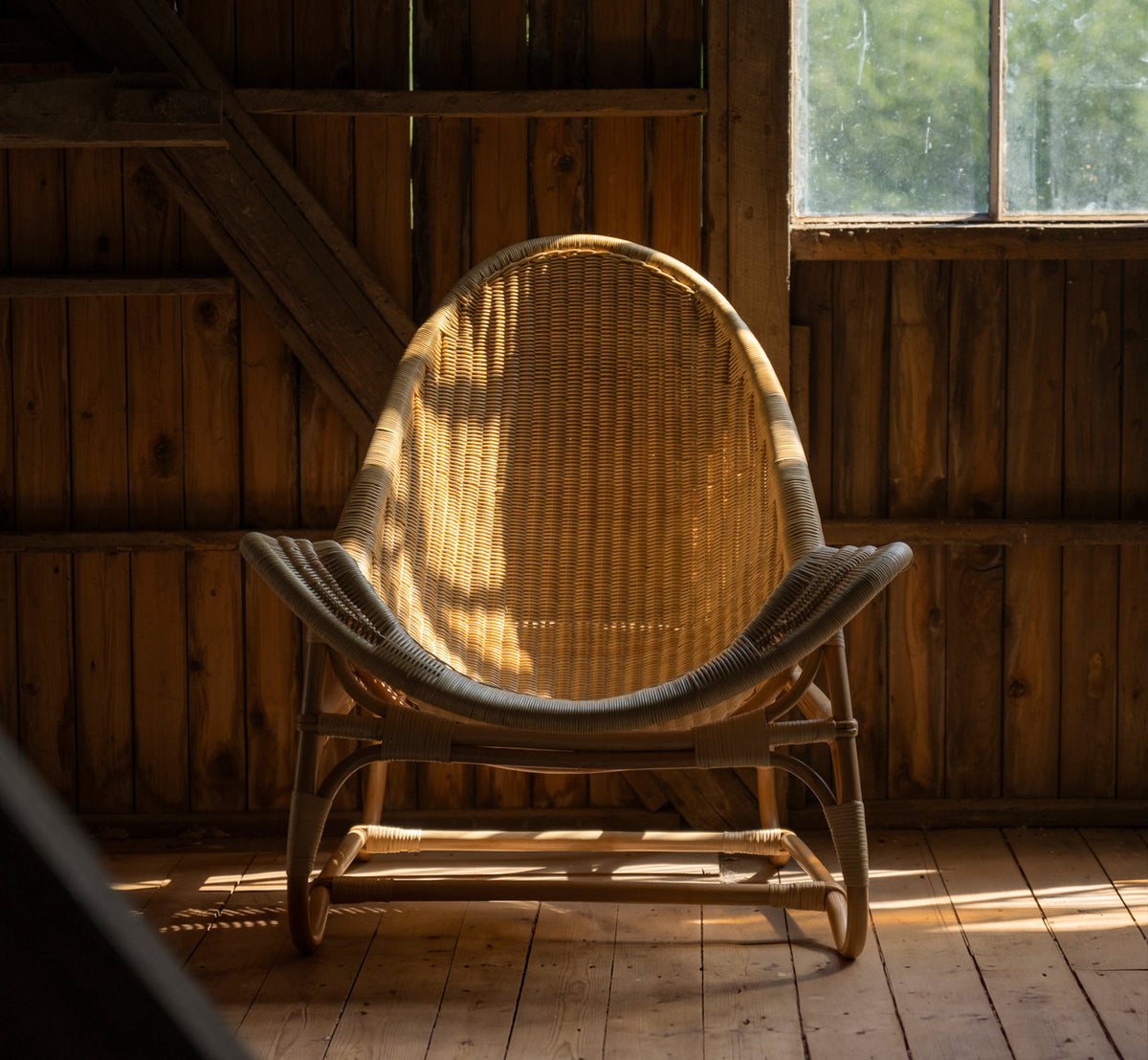 Dette er et livsstilsbillede af Pacifique Lænestol i natural core weave rattan frame. I en træklædt lade med varmt lys fra vinduet.