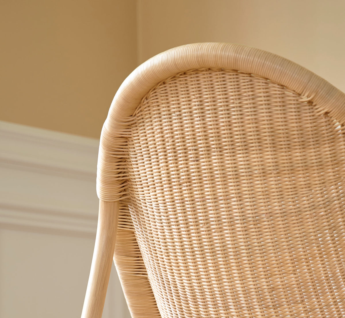 Dette er et livsstilsbillede af Pacifique Lænestol i natural core weave rattan frame. Flettet ryg i blødt lys ved en panelvæg.