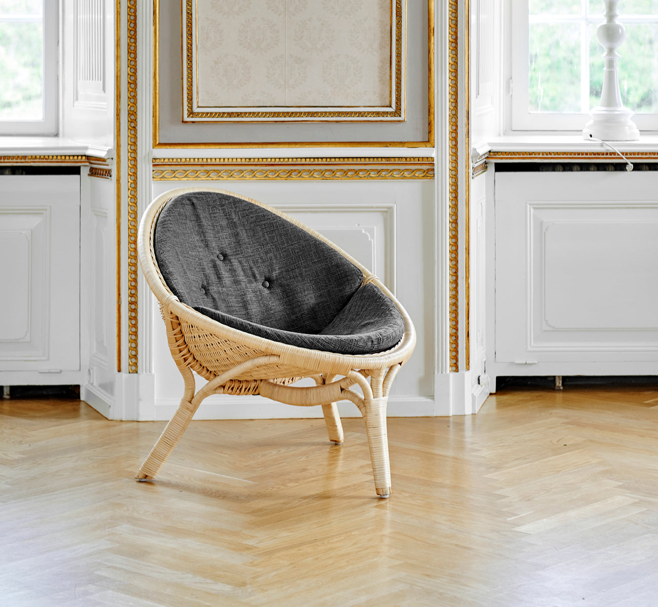 Dette er et livsstilsbillede af Rana Lænestol i natural core weave rattan frame. Stående alene i et rum med forgyldt panel og parketgulv.
