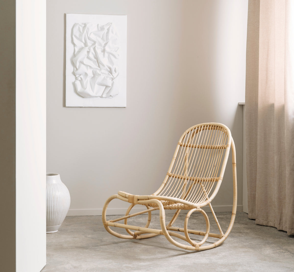 Dette er et livsstilsbillede af Nanny Rocking Chair i polished natural rattan. I en galleriopsætning med andre rattanmøbler på piedestaler.