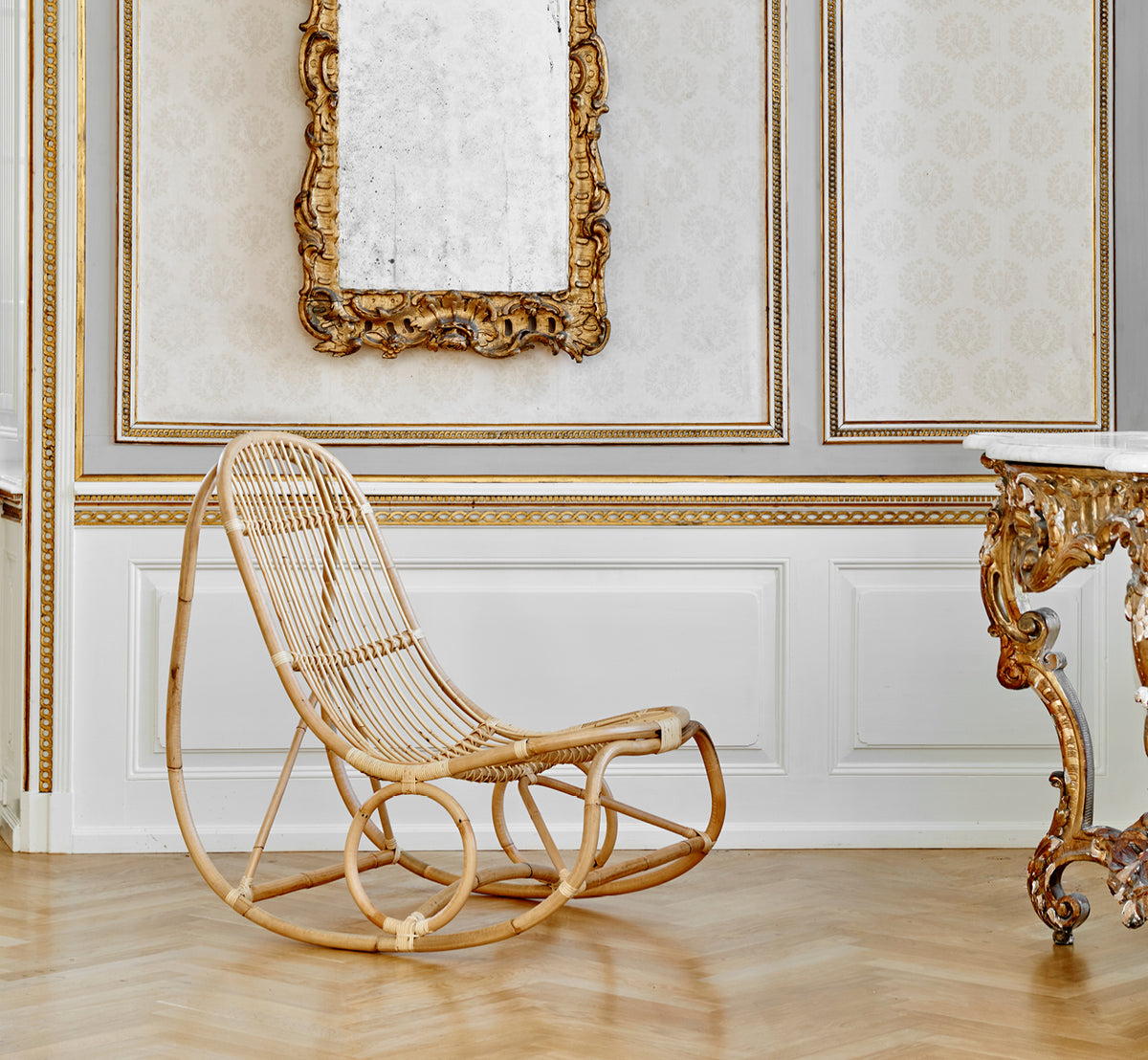 Dette er et livsstilsbillede af Nanny Rocking Chair i polished natural rattan. I et udsmykket forgyldt rum med barokspejl og konsol.