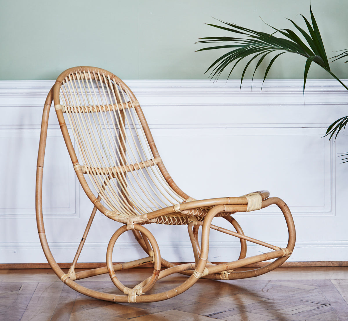 Dette er et livsstilsbillede af Nanny Rocking Chair i polished natural rattan. Mod en salviefarvet grøn væg med hvide paneler og palme.