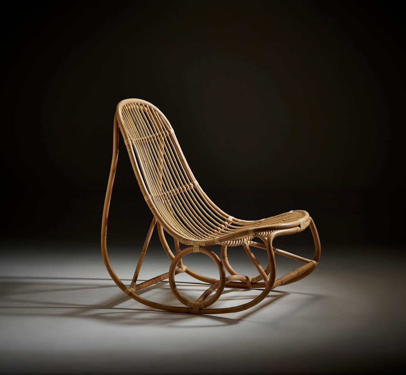 Dette er et livsstilsbillede af Nanny Rocking Chair i polished natural rattan. Studiefoto på mørk baggrund, der fremhæver formerne.