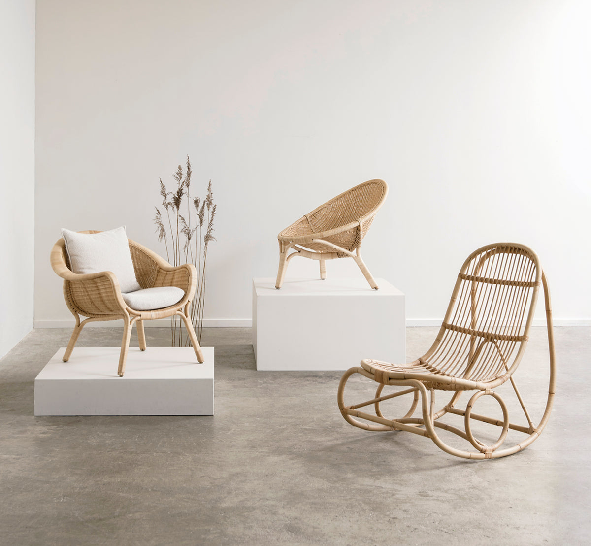 Dette er et livsstilsbillede af Nanny Rocking Chair i polished natural rattan. I et galleri med rattanstole på hvide udstillingspiedestaler.