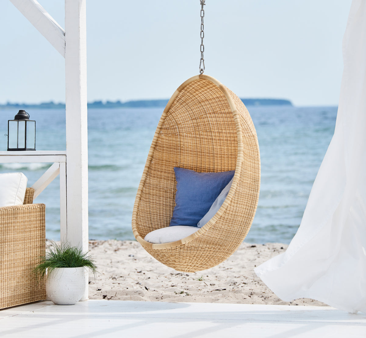 Dette er et livsstilsbillede af Hanging Udendørs hængeæg-stol i natural core weave ArtFibre Aluminium, hængende fra en hvid pergola med sandstrand og.