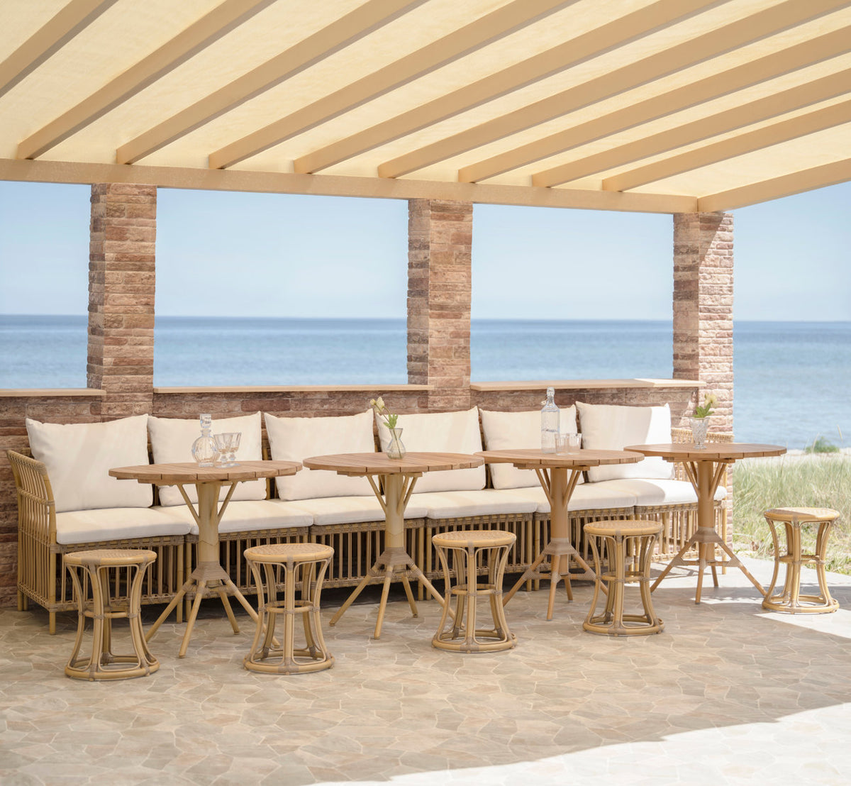Dette er et livsstilsbillede af Meghan Udendørs spisebænk-Center Module i natural aluminium artfibre. Under en pergola ved havet.