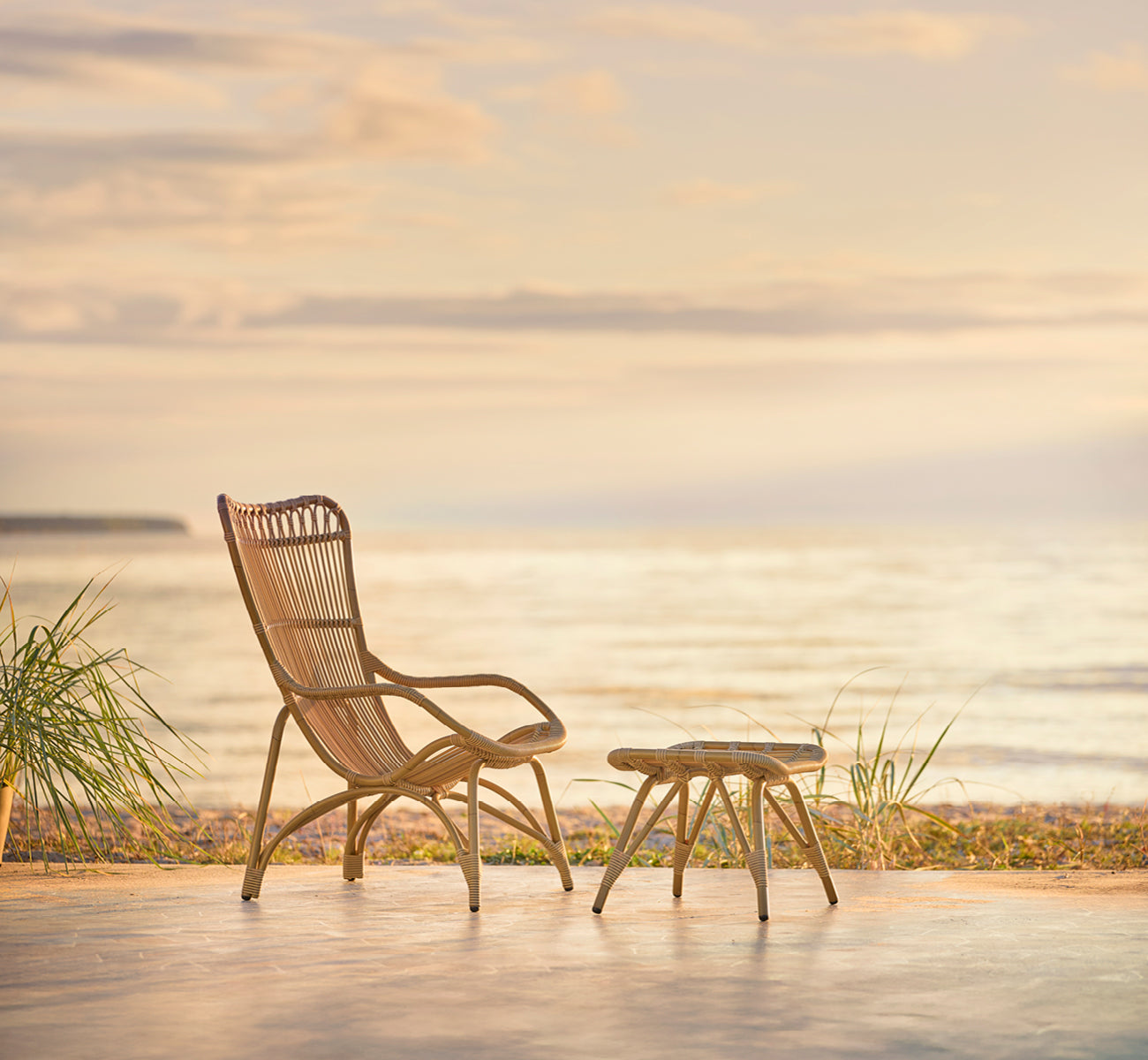 Dette er et livsstilsbillede af Monet Exterior Footstool i antique aluminium artfibre. Med lænestol på et dæk i en strandpavillon.