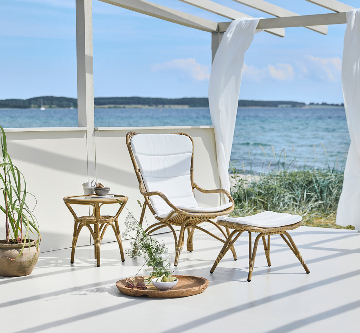 Dette er et livsstilsbillede af Monet Exterior Footstool i antique aluminium artfibre. I en strandpavillon med polstret stol.