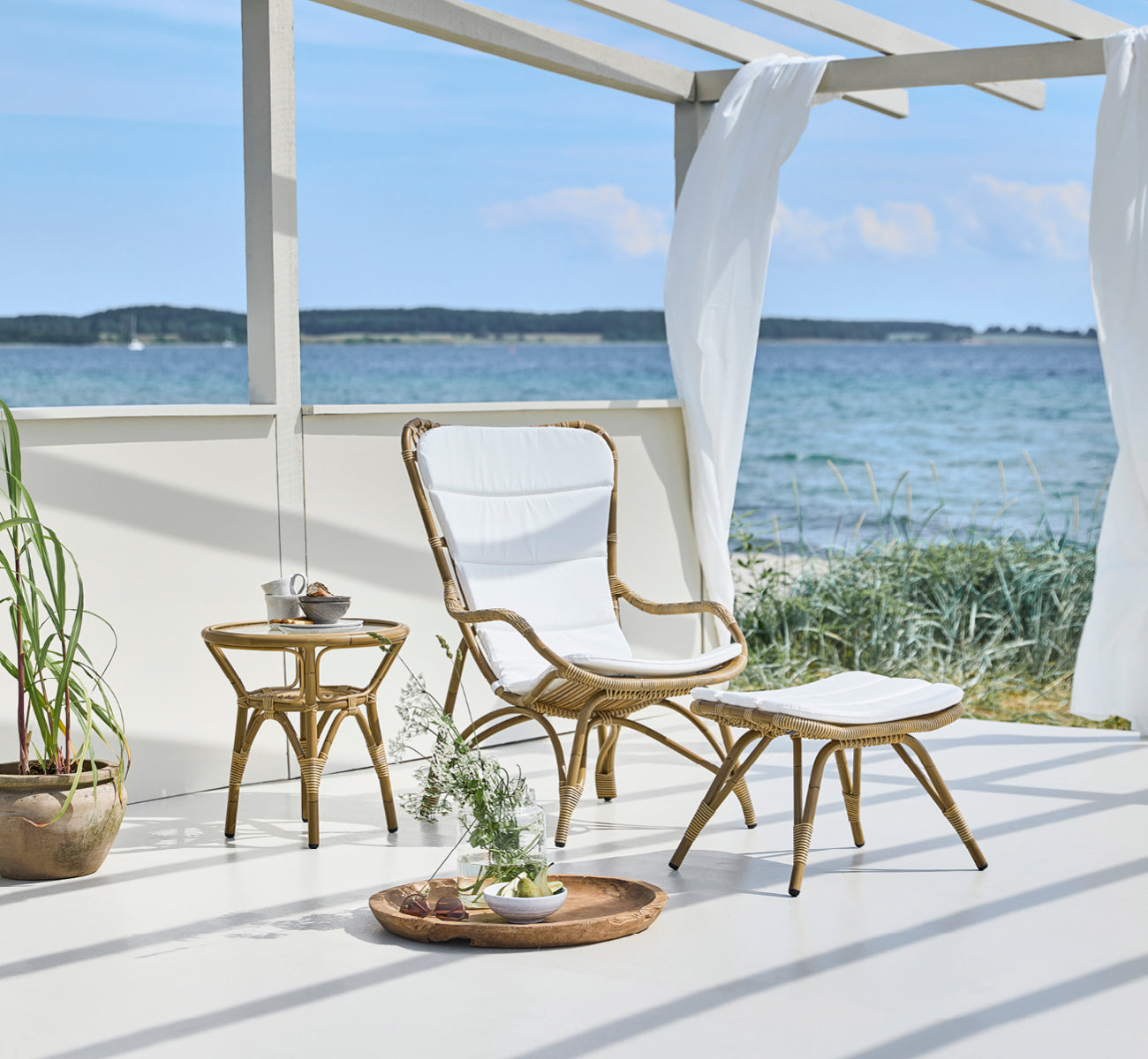 Dette er et livsstilsbillede af Monet Exterior Footstool i antique aluminium artfibre. I en strandpavillon med polstret stol.