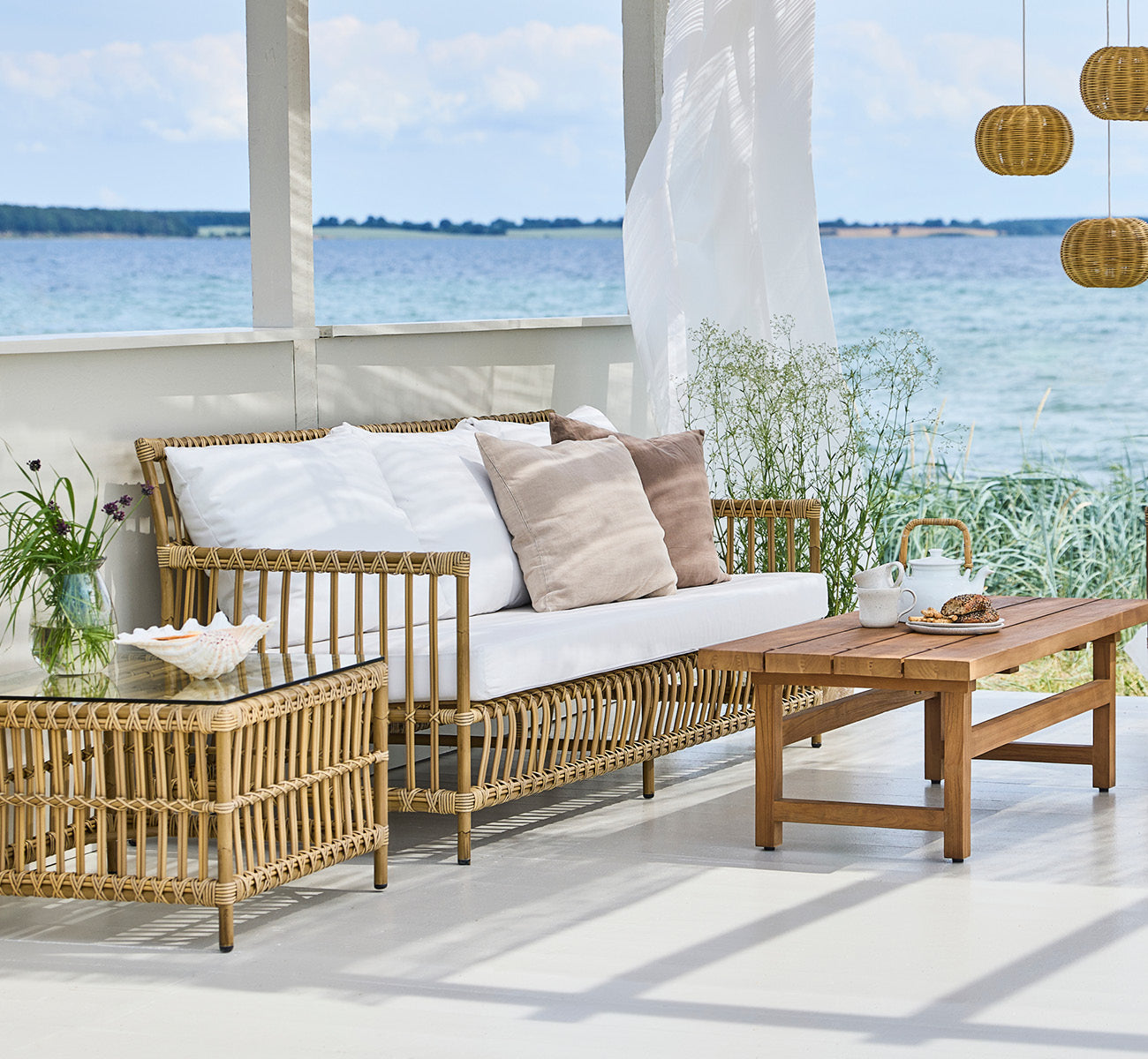 Dette er et livsstilsbillede af Caroline Udendørs sofa i antique Aluminium ArtFibre, på et trædæk ved vandet med kystnær vegetation og havudsigt.