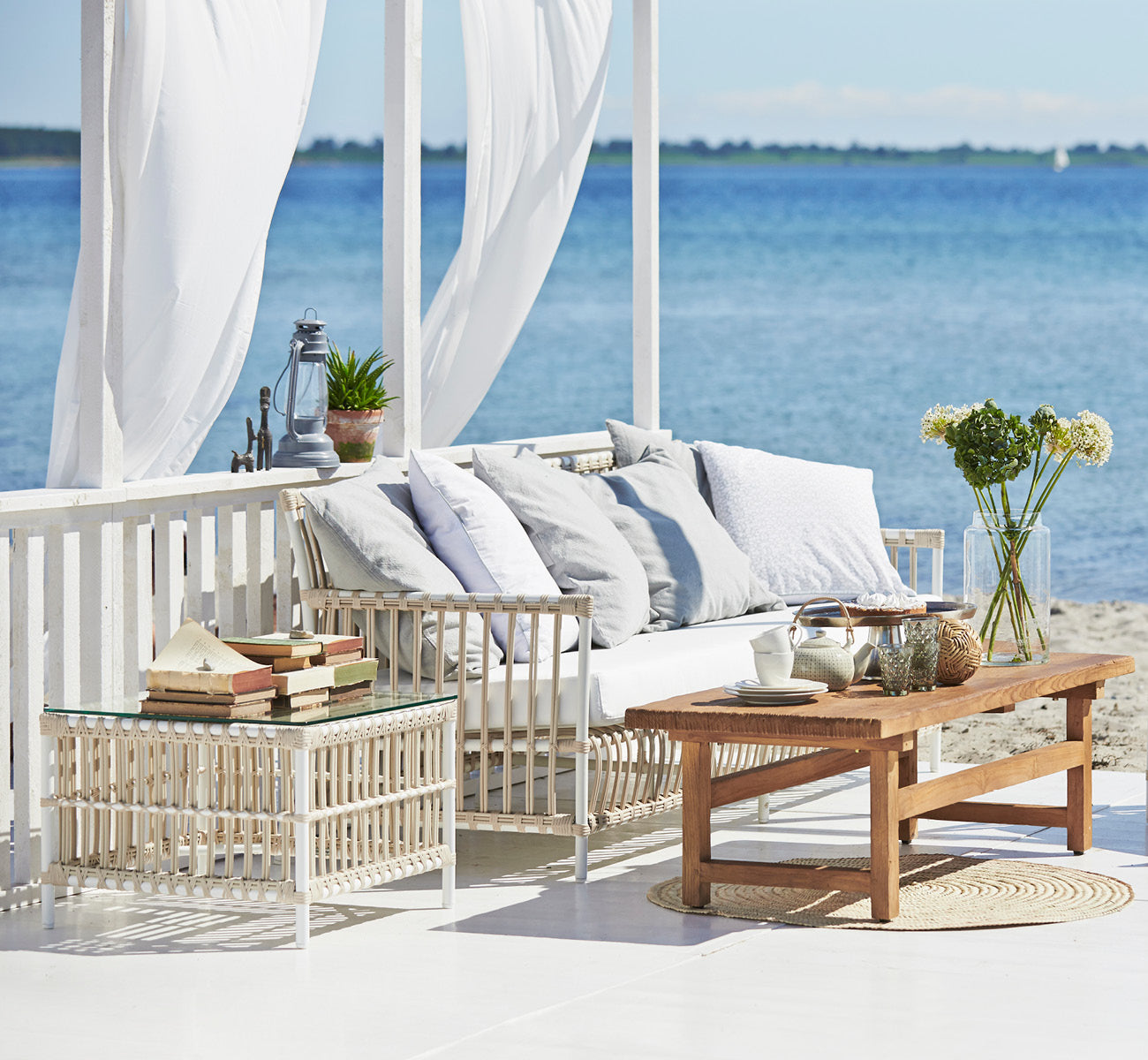 Dette er et livsstilsbillede af Caroline Udendørs sofa i dove White Aluminium ArtFibre, på et trædæk ved vandet med kystnær vegetation og havudsigt.