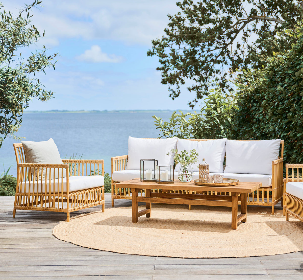 Dette er et livsstilsbillede af Caroline Udendørs sofa i natural Aluminium ArtFibre, på et trædæk ved vandet med kystnær vegetation og havudsigt.