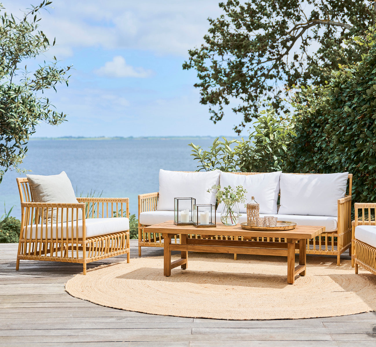 Dette er et livsstilsbillede af Caroline Udendørs sofa i natural Aluminium ArtFibre, på et trædæk ved vandet med kystnær vegetation og havudsigt.