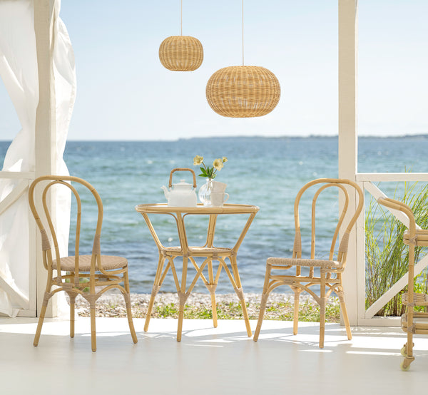 Dette er et livsstilsbillede af Tony Udendørs cafébord i natural aluminium artfibre. På en strandterrasse med stole, pendellampe og havudsigt.