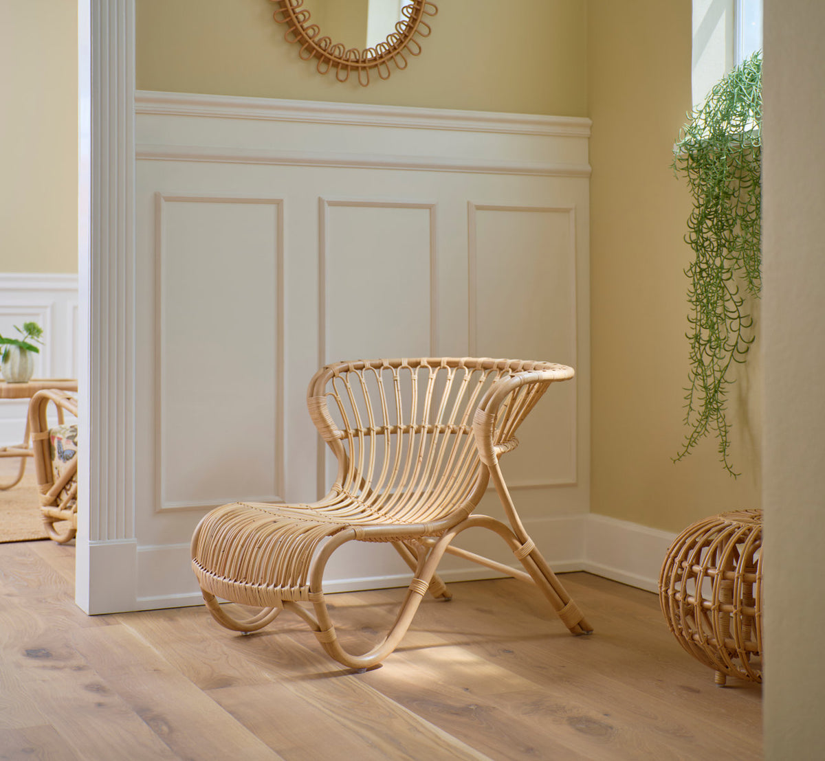 Dette er et livsstilsbillede af Fox Lænestol i polished natural rattan. Ved hvide paneler med et rattanspejl.