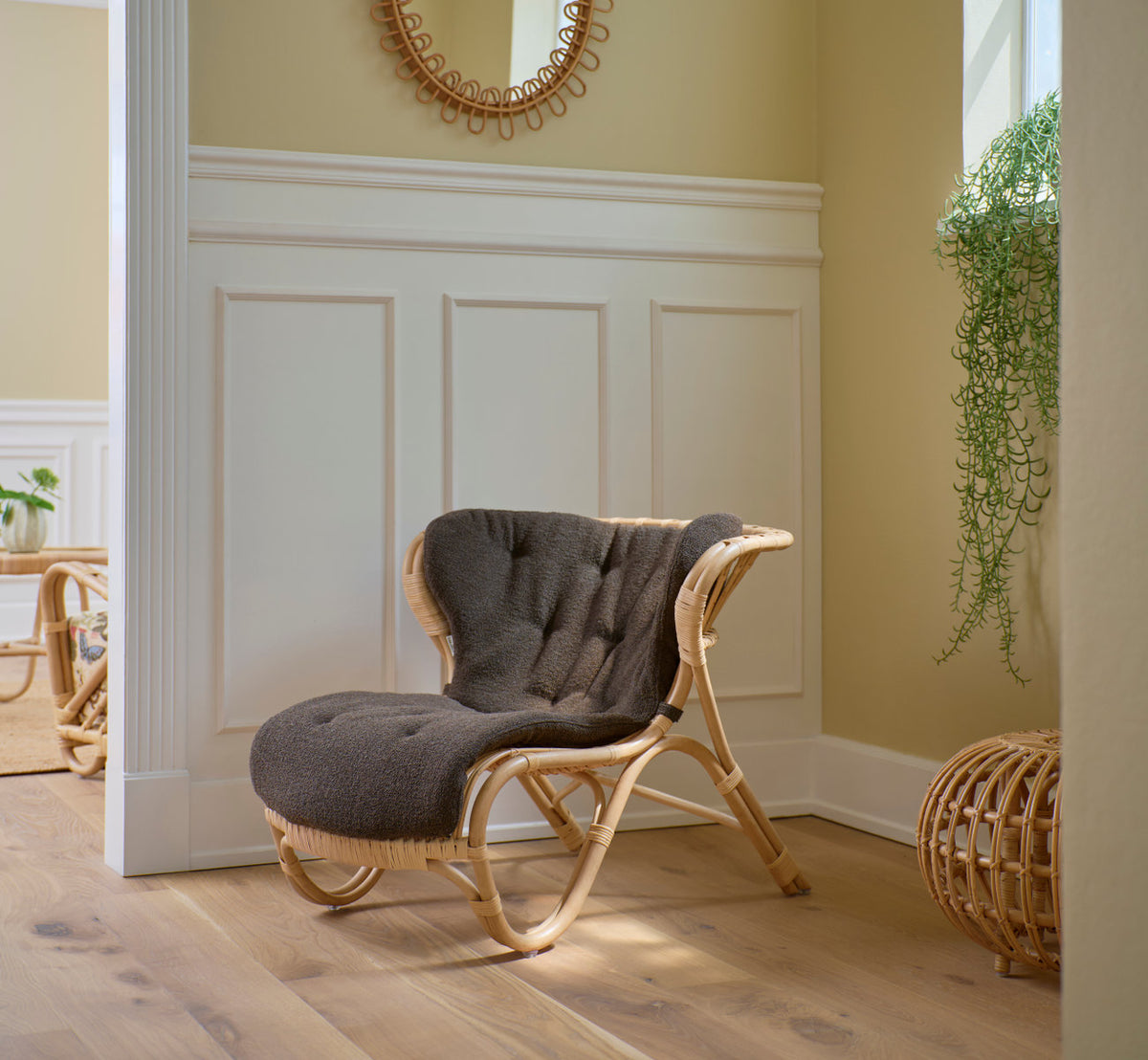 Dette er et livsstilsbillede af Fox Lænestol i polished natural rattan. Med grå pude ved hvide paneler.