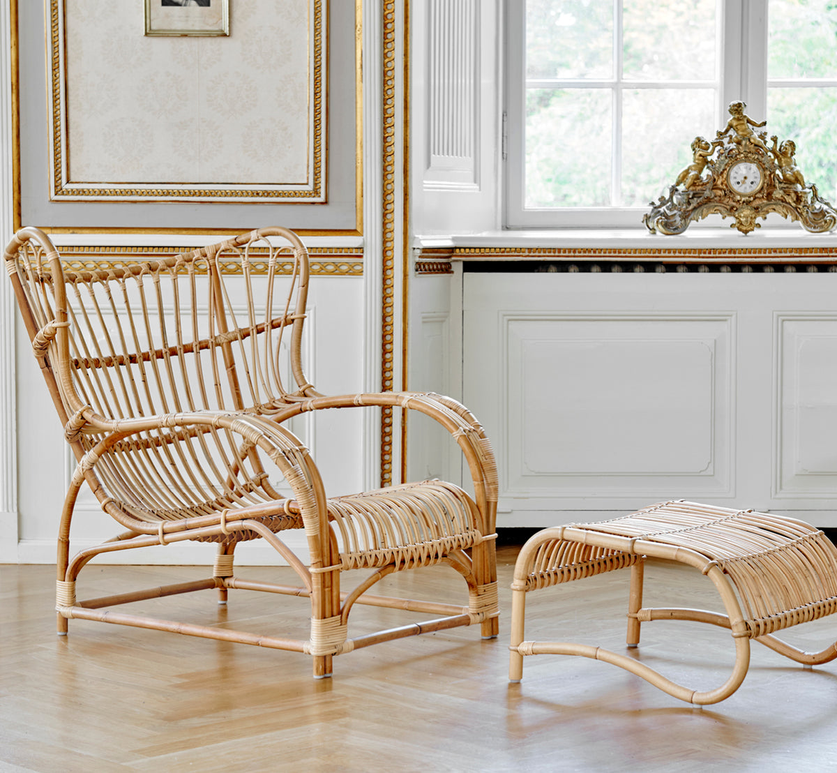 Dette er et livsstilsbillede af Teddy Fodskammel i polished natural rattan. Kombineret med den matchende lænestol ved et lyst gardinvindue.