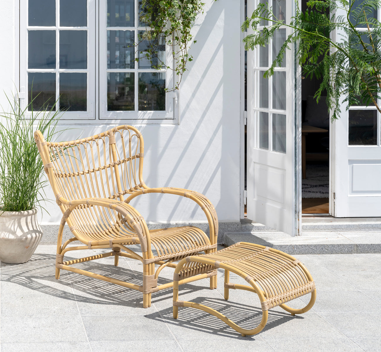 Dette er et livsstilsbillede af Teddy Udendørs fodskammel i natural aluminium artfibre. Kombineret med den matchende lænestol på en solbeskinnet brolagt terrasse.