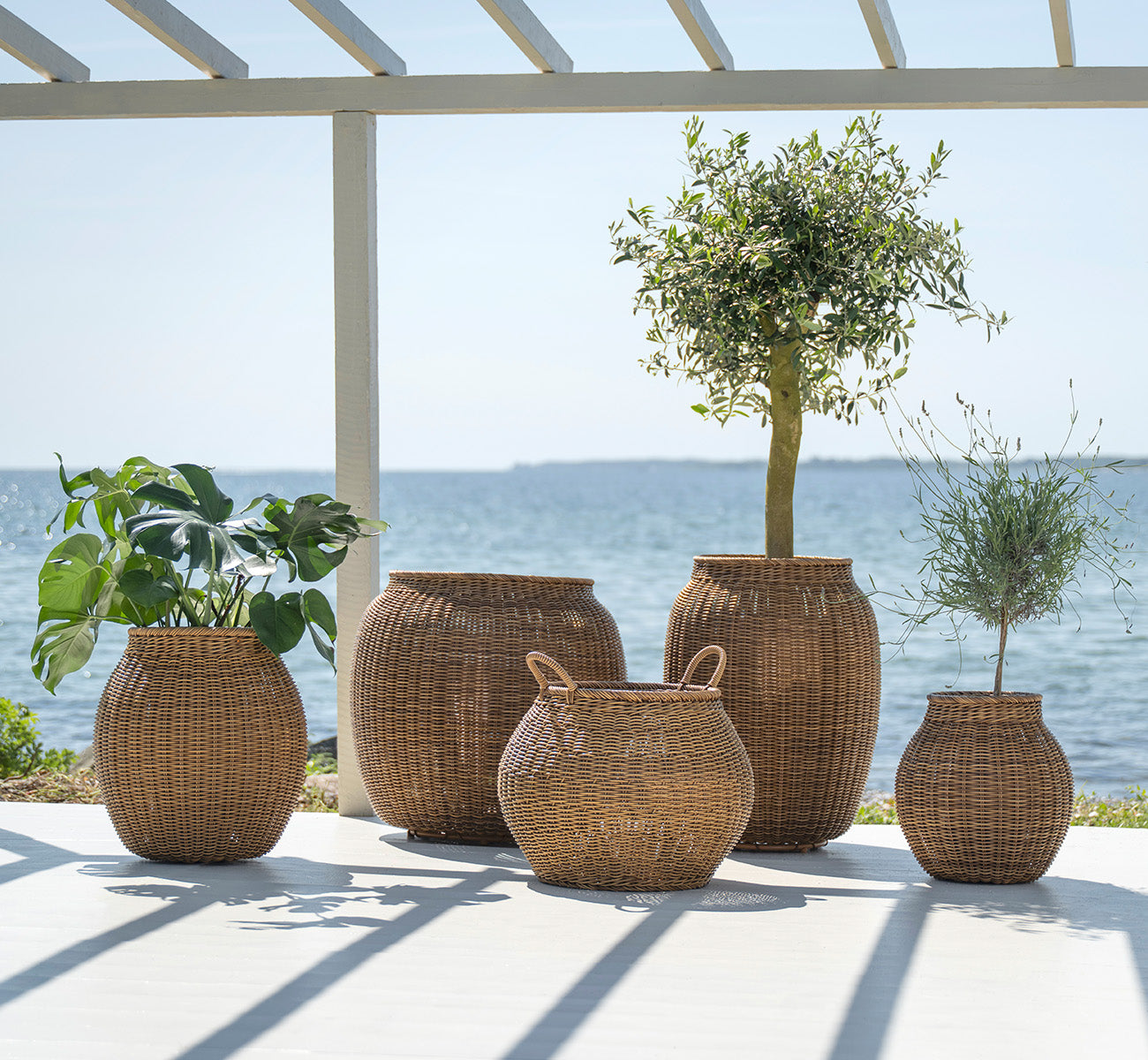 Dette er et livsstilsbillede af Alma Kurv i chestnut core weave ArtFibre Aluminium, udendørs på en solbeskint terrasse med hvid mur og flisegulv.