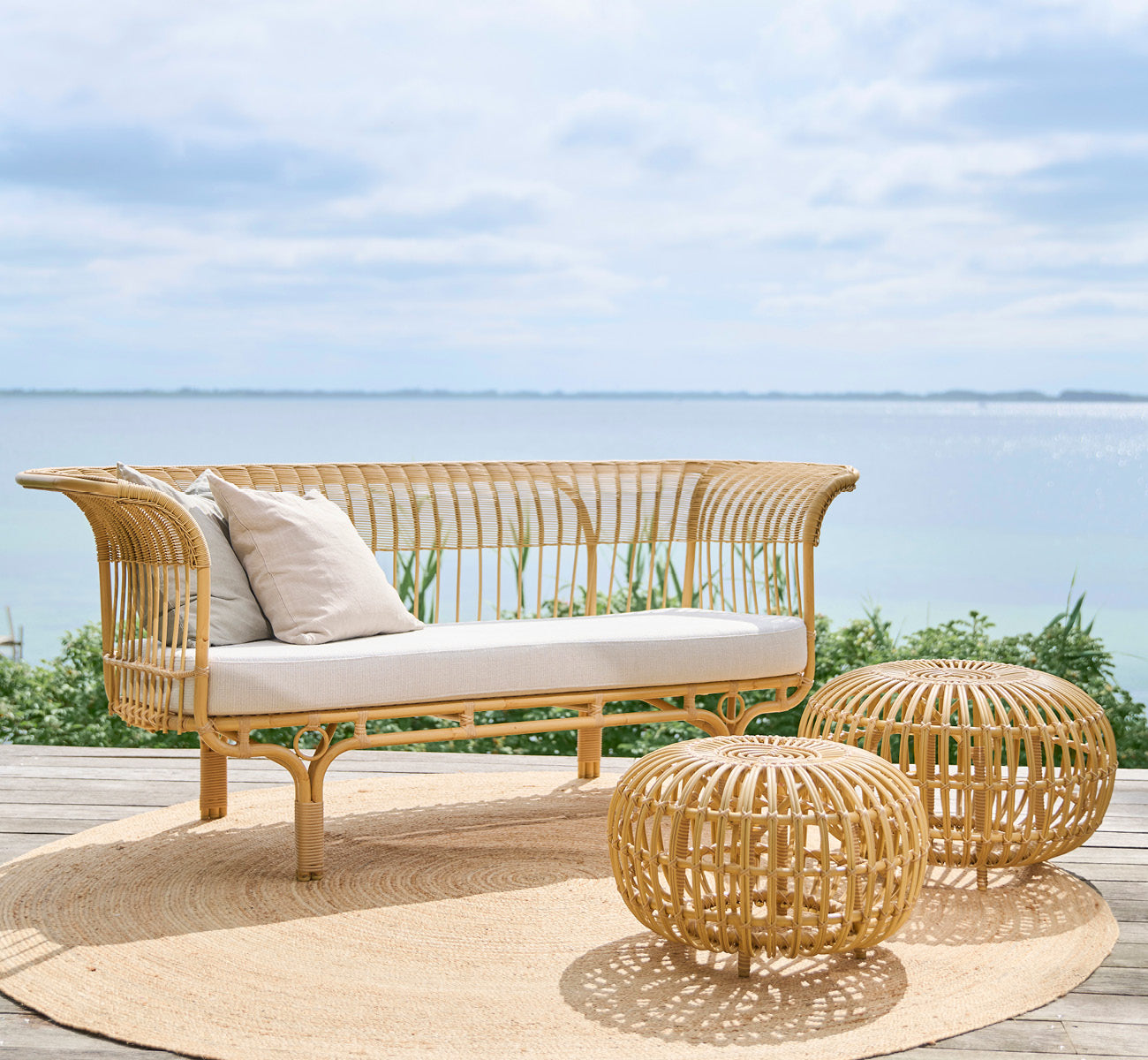 Dette er et livsstilsbillede af Belladonna Udendørs sofa i natural Aluminium ArtFibre, på et udendørs trædæk med udsigt over vandet.