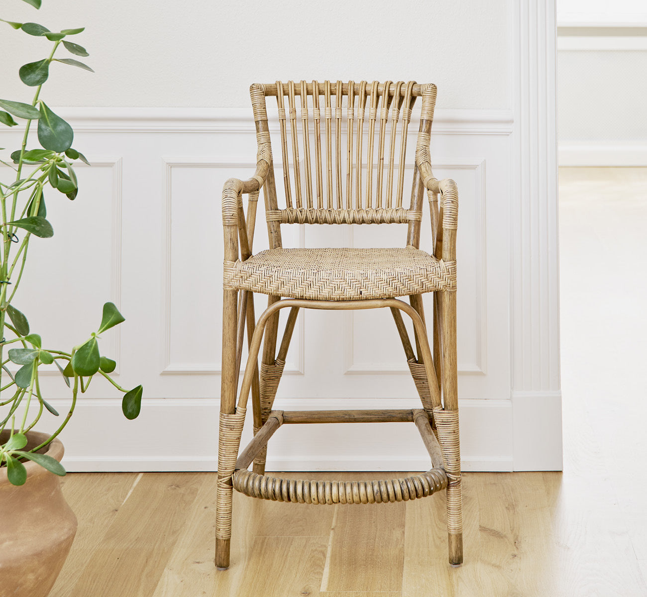Dette er et livsstilsbillede af Blues Skammel i antique Rattan, i et lyst interiør med hvide panelvægge og sildebensgulv.
