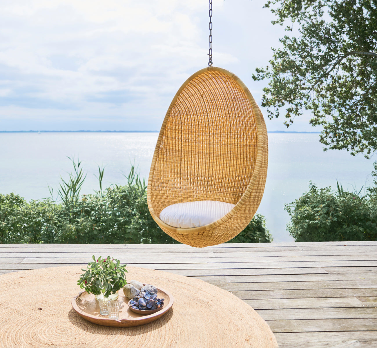 Dette er et livsstilsbillede af Hanging Udendørs hængeæg-stol i natural core weave ArtFibre Aluminium, hængende fra en hvid pergola med sandstrand og.