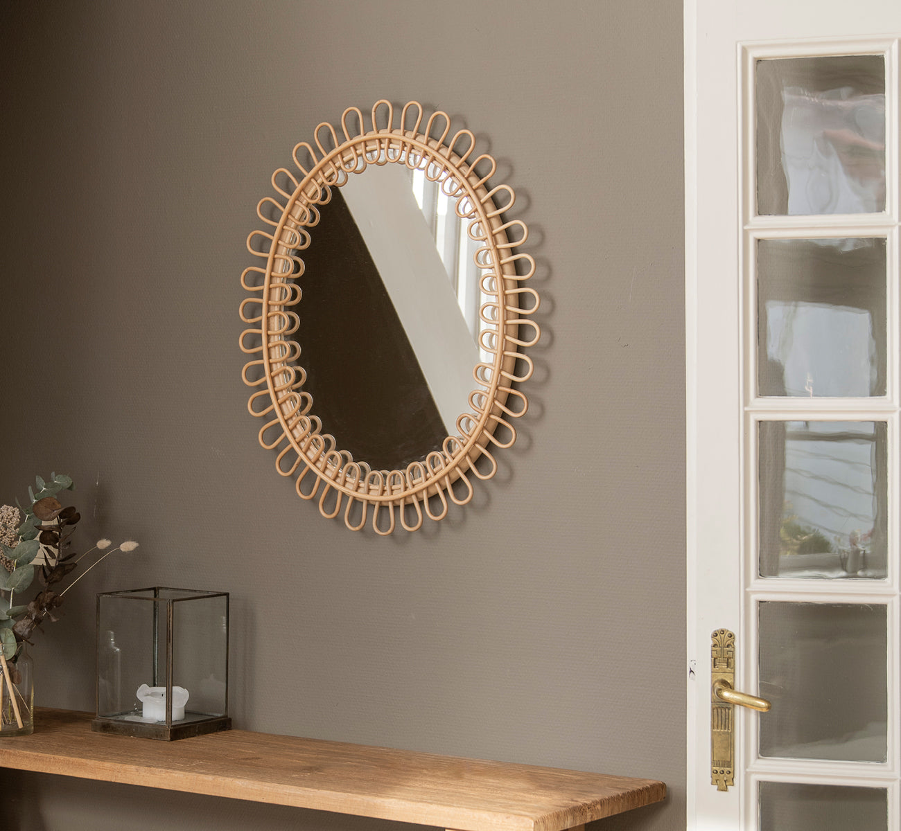 Dette er et livsstilsbillede af Luella Spejl i polished natural rattan. Hængende på en grå væg over en træhylde med eukalyptus og glasdekoration.