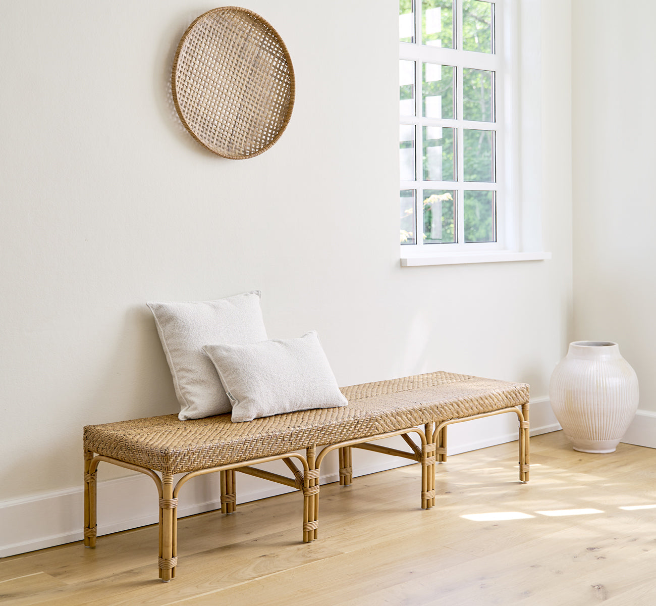 Dette er et livsstilsbillede af Luis Bænk i antique rattan. Stylet med linnepuder i et lyst hvidt rum med vægdekoration og en hvid vase.
