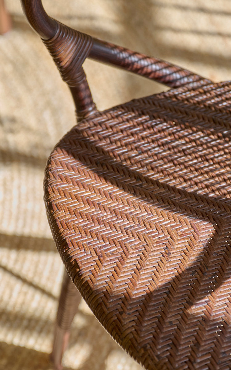 Dette er et nærbillede af Rossini Spisestuelænestol i brown rattan. Sildebensflettet sæde og omviklet armlæn i varmt sollys.