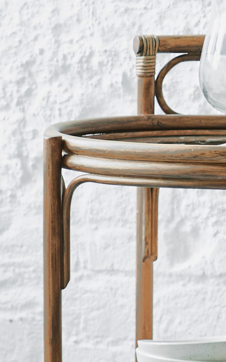 Dette er et nærbillede af Romeo Rullebord i antique rattan. Detalje af det runde rattan-rullebordsstel mod en hvid murstensvæg.