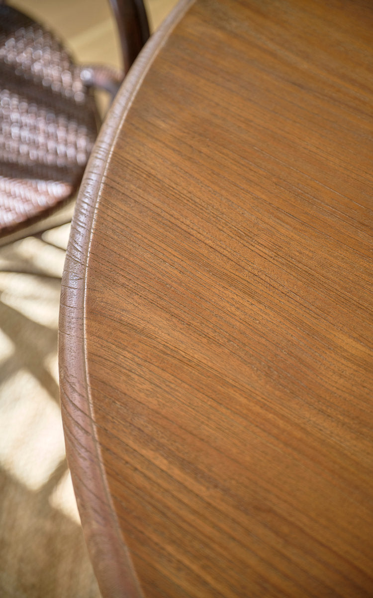 Dette er et nærbillede af Michel Teak spisebord Ø120 cm i teak indoor. Kant af rund teakbordplade med rattanstol bagved.