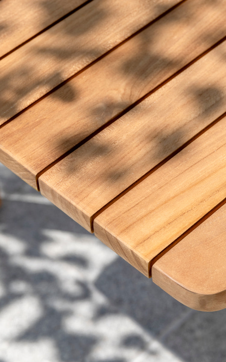 Dette er et nærbillede af Tobias Exterior Teak Table 205x91 cm i teak outdoor. Detalje af teakplankebordpladekanten og plettet sollys.