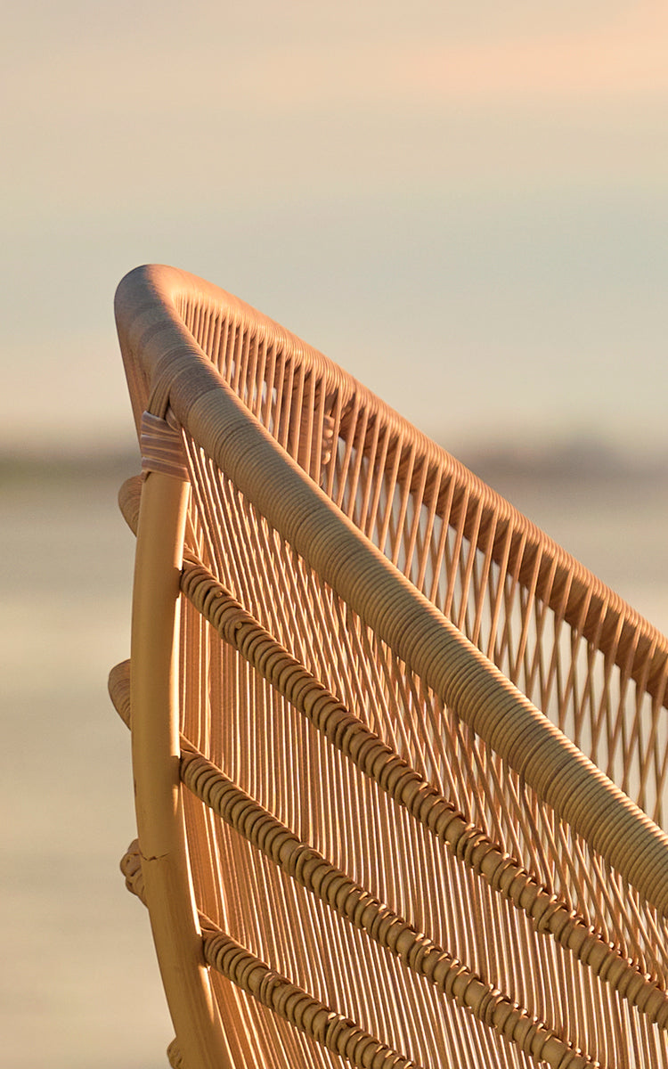 Dette er et nærbillede af Luna Udendørs lænestol i natural aluminium artfibre. Det flettede ryglæn lyser op i gyldent solnedgangslys ved havet.
