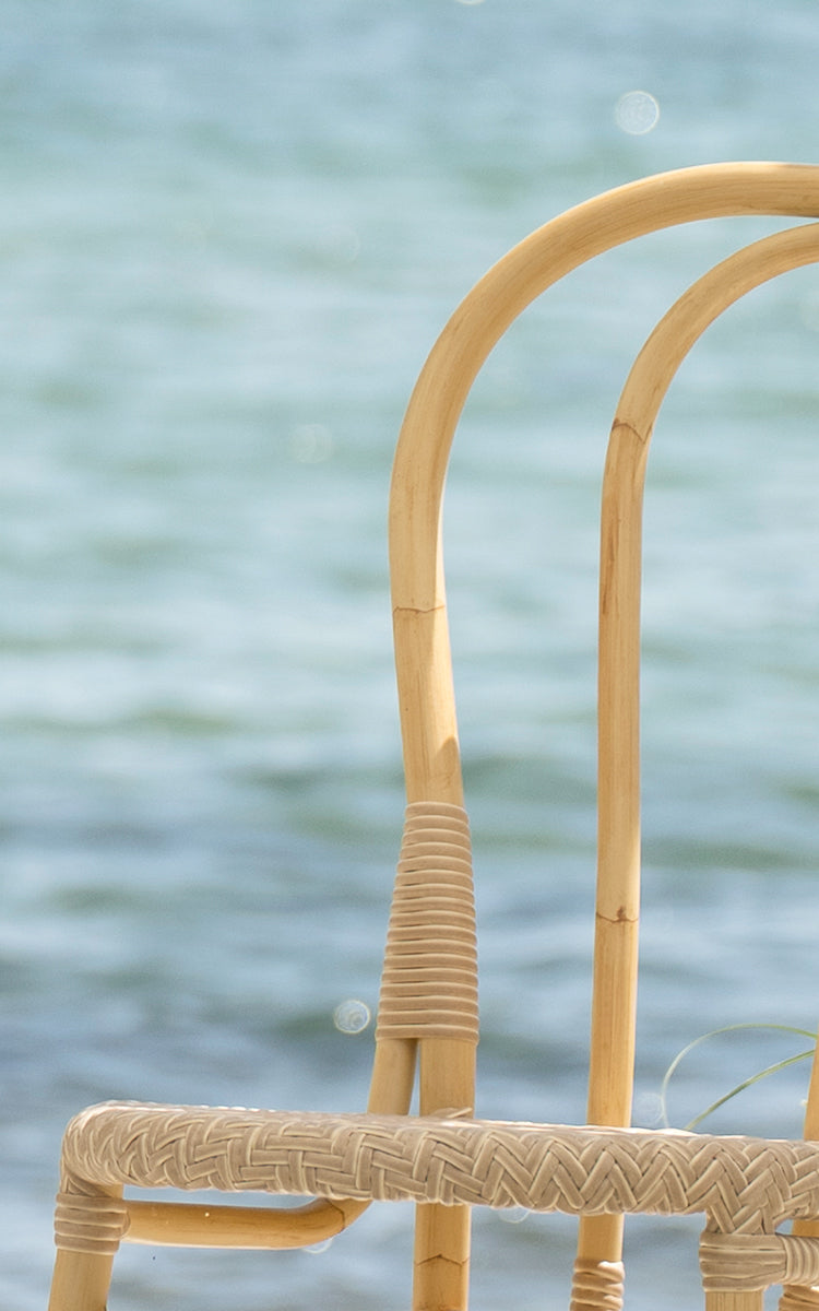Dette er et nærbillede af Lulu Udendørs spisebordsstol i natural aluminium artfibre. Det buede stel og den flettede detalje er vist ved havet.