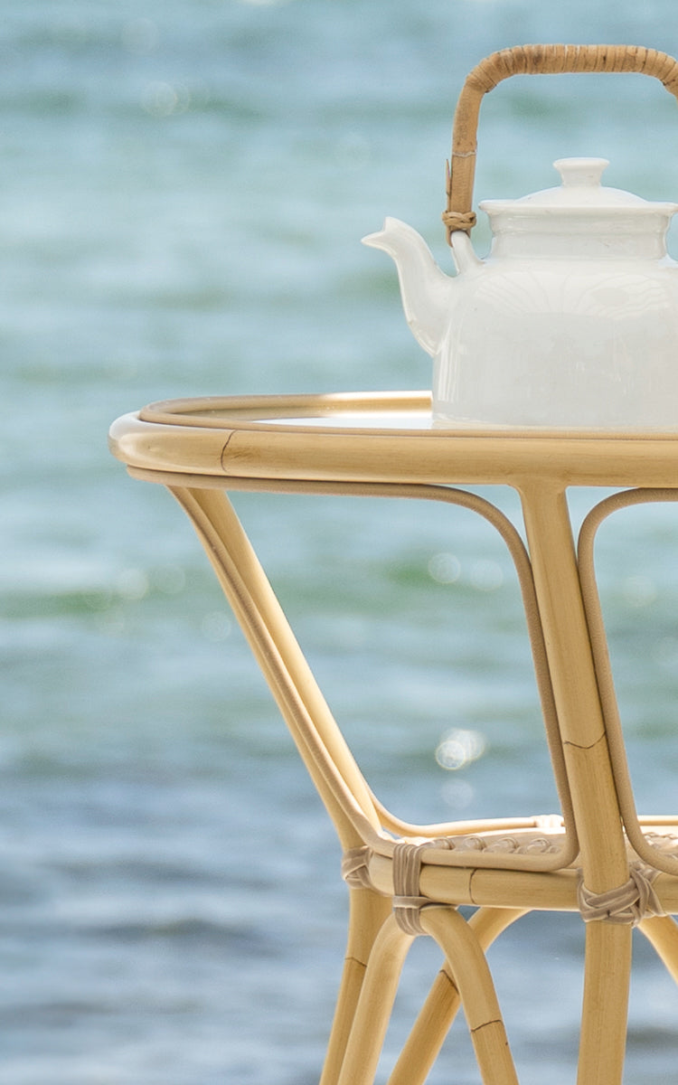 Dette er et nærbillede af Tony Udendørs cafébord i natural aluminium artfibre. En hvid tekande står på den runde bordplade med havet i baggrunden.
