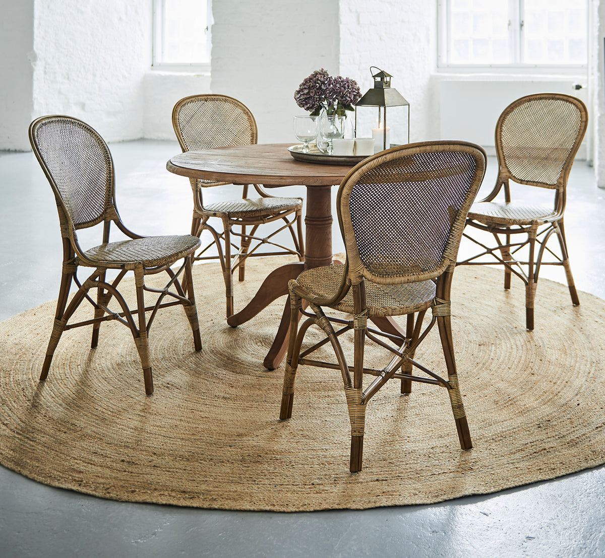 Dette er et livsstilsbillede af Rossini Dining Chair i antique rattan. Fire stole omkring et rundt teakbord på et jutetæppe i en hvid loft.