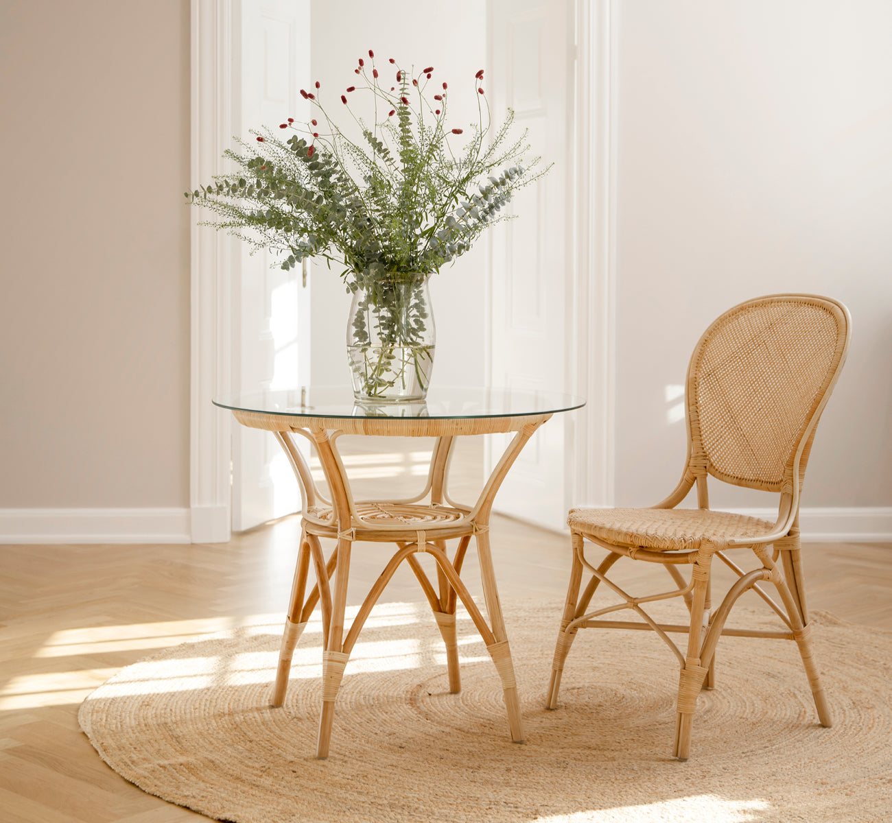 Dette er et livsstilsbillede af Rossini Spisebordsstol i natural rattan. Placeret ved siden af et bistrobord med glasplade på et jutetæppe i et lyst hallway.
