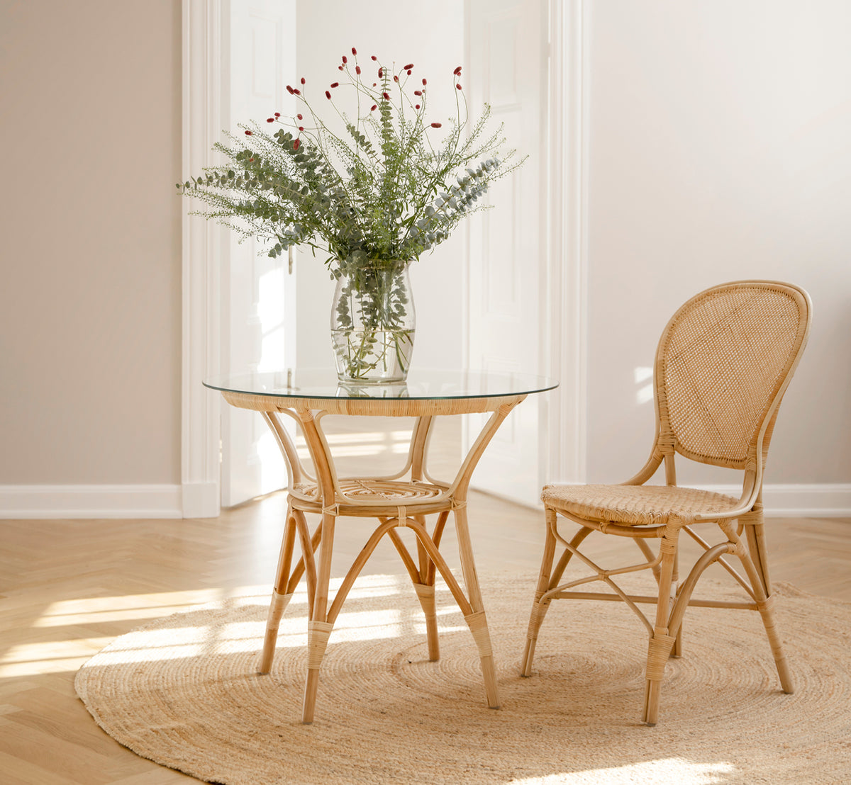 Dette er et livsstilsbillede af Rossini Spisebordsstol i natural rattan. Placeret ved siden af et bistrobord med glasplade på et jutetæppe i et lyst hallway.