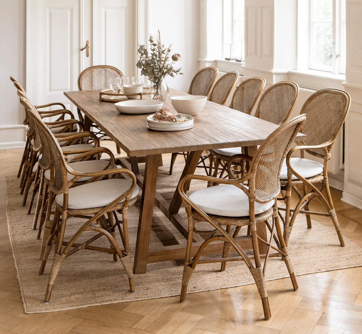 Dette er et livsstilsbillede af Rossini Spisestuelænestol i antique rattan. Omkring et langt teakbord med lindrapering på et rundt jutetæppe.