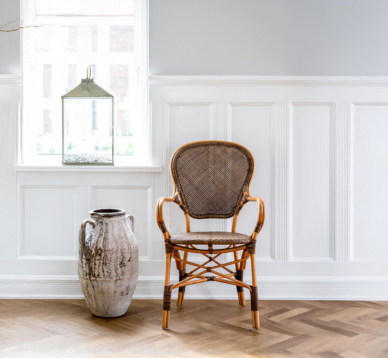 Dette er et livsstilsbillede af Rossini Spisestuelænestol i cherry rattan. Opstillet i et panelrum med sildebensgulv ved siden af en rustik urne.
