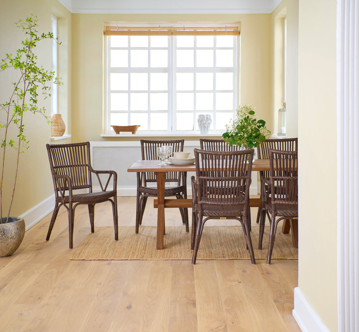 Dette er et livsstilsbillede af Piano Spisebordsstol i brown rattan. Omkring et teak spisebord i et lyst gult rum.