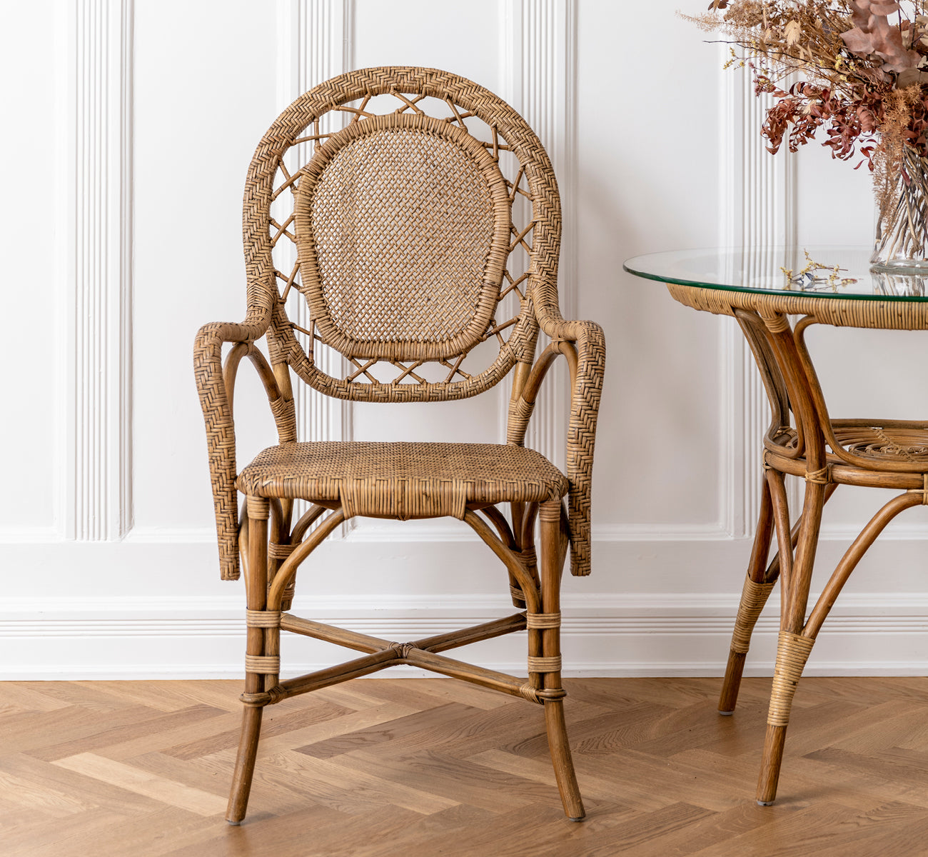 Dette er et livsstilsbillede af Romantica Spisebordsstol i antique rattan. Placeret ved et glasbord i rattan med tørrede blomster i et panelrum.