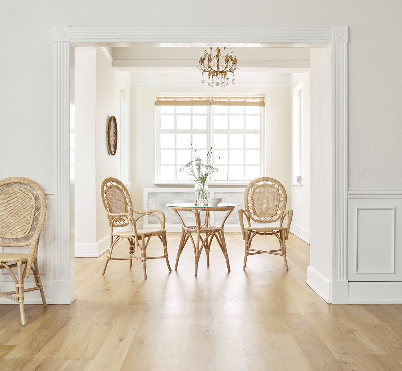 Dette er et livsstilsbillede af Romantica Spisebordsstol i natural rattan. Omkring et rundt bord i et elegant rum med lysekrone og hvide paneler.