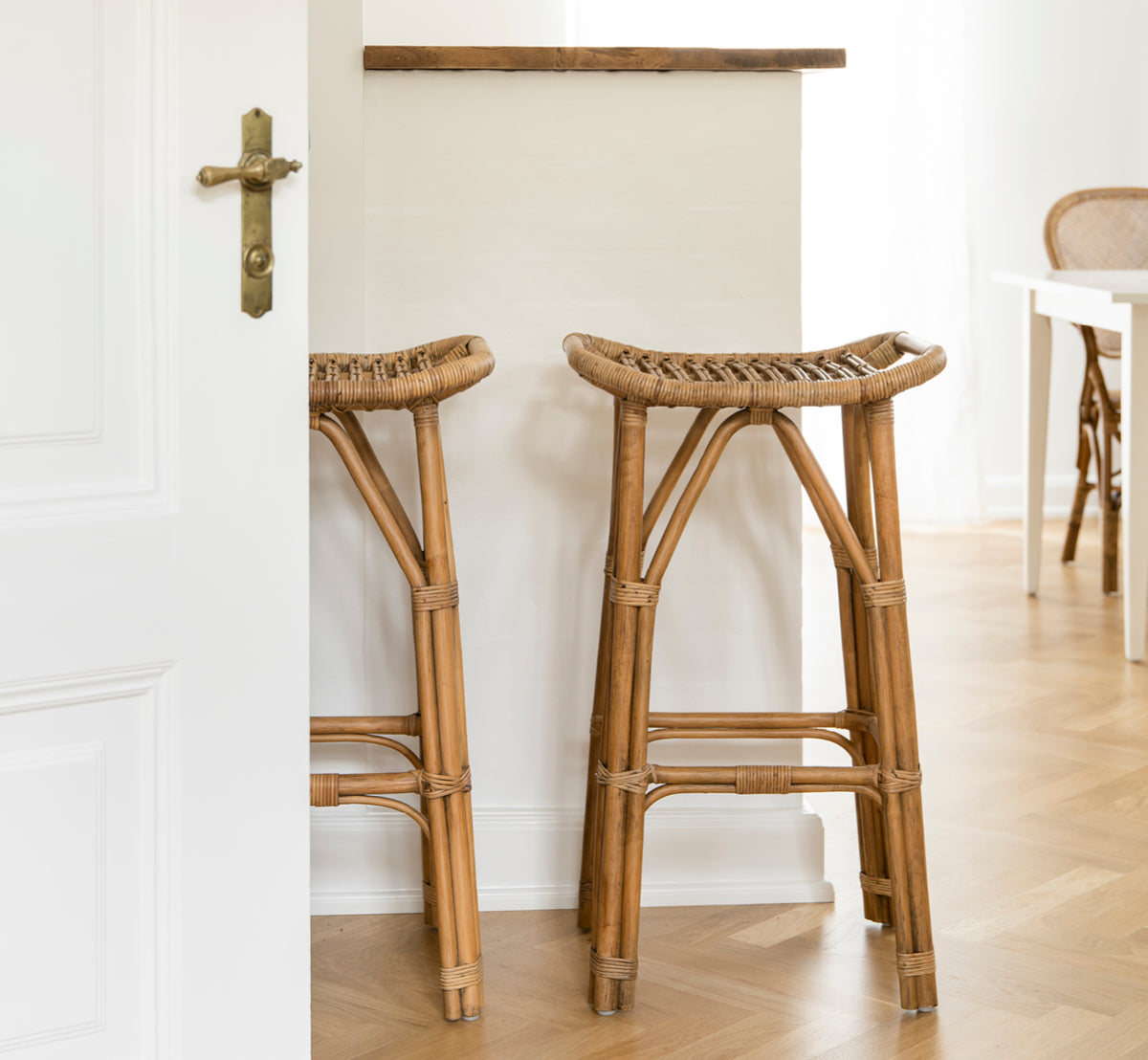 Dette er et livsstilsbillede af Salsa Barstol i antique rattan. Et par placeret mod en hvid væg på et sildebensparketgulv.