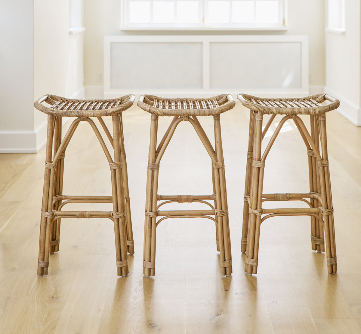Dette er et livsstilsbillede af Salsa Barstol i antique rattan. Et par placeret mod en hvid væg på et sildebensparketgulv.