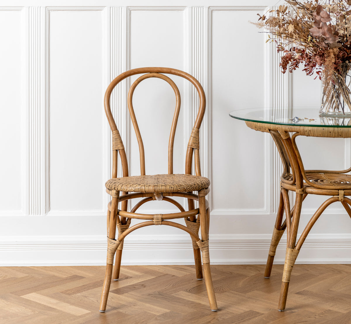 Dette er et livsstilsbillede af Lulu Spisebordsstol i antique rattan. Placeret ved et glasbord i rattan med tørrede blomster i et panelinteriør.