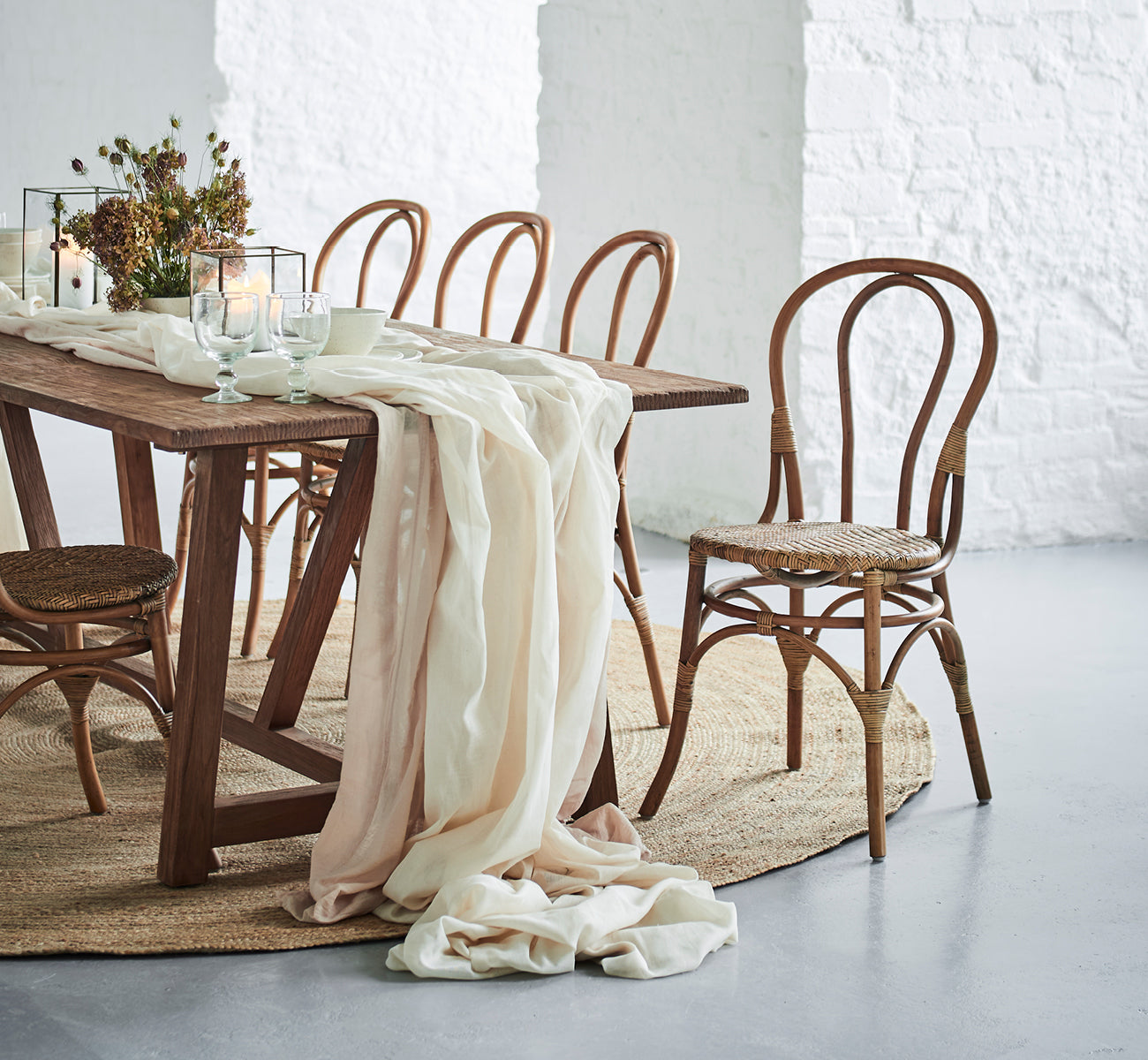 Dette er et livsstilsbillede af Lulu Spisebordsstol i antique rattan. Placeret ved et rustikt bord med lindrapering og stearinlys mod en hvid murstensvæg.