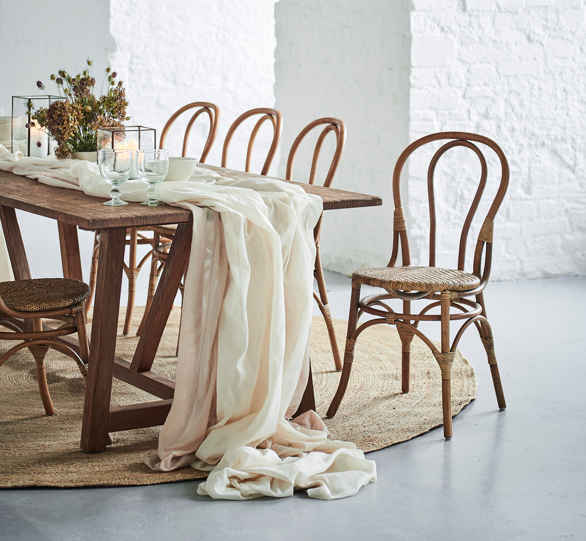 Dette er et livsstilsbillede af Lulu Spisebordsstol i antique rattan. Placeret ved et rustikt bord med lindrapering og stearinlys mod en hvid murstensvæg.